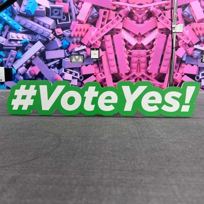 Green and white hashtag cutout sign with the text #VoteYes! against a colorful Lego brick background.