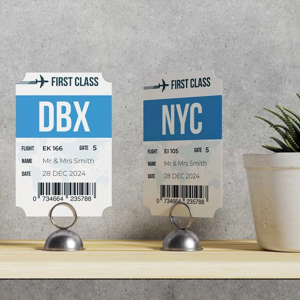 Two wedding table name cards designed as first class boarding passes, showing airport codes DBX and NYC with couple names and wedding date. Printed on rigid board in silver metal card holders.
