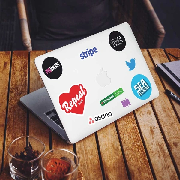 A MacBook on a wooden table covered in die-cut laptop stickers. Visible logos include Stripe, Hozier, Sea Sessions, Pygmalion, Asana, Twitter, Heineken Music, and a Repeal sticker. 
