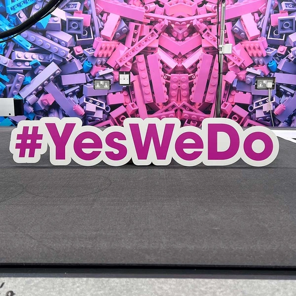 Pink and white hashtag cutout sign with the text #YesWeDo against a vibrant Lego brick background.